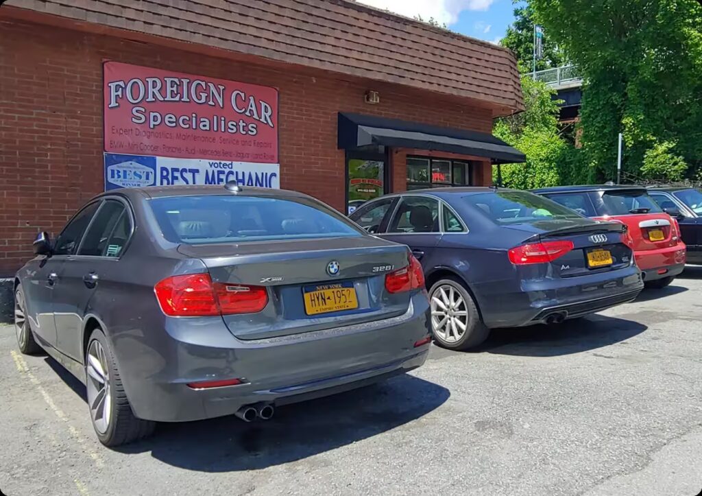 Two cars, a BMW and an Audi, are parked outside a brick building with a sign that reads Foreign Car Specialists. The license plates are visible, and the background includes trees and a partly cloudy sky.