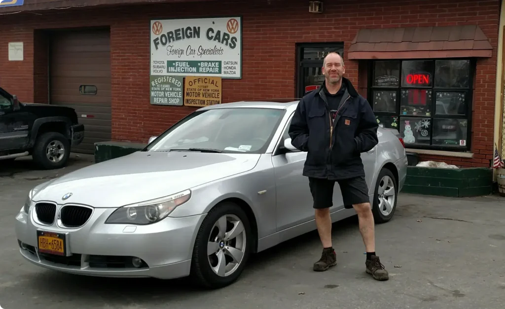 A man wearing a dark jacket and shorts stands smiling next to a silver BMW sedan. The car is parked in front of a brick building with a sign that reads Foreign Cars and various service offerings. An Open sign is visible in the window.