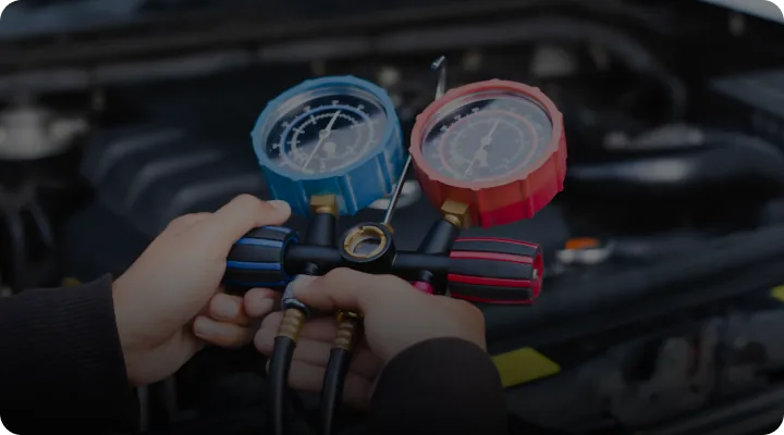 Hands holding a set of HVAC manifold gauges with blue and red dials, used for measuring pressure in an air conditioning system, with a car engine in the background.