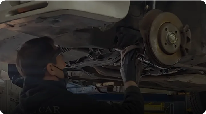 A mechanic wearing a mask and gloves is working underneath a lifted car. The vehicles wheel and brake disc are visible. The mechanic appears focused on inspecting or repairing part of the cars undercarriage.