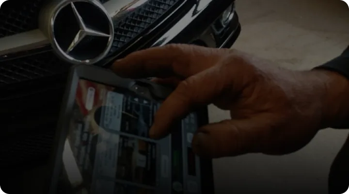 Close-up of a hand holding a tablet with a blurred screen, in front of a black car featuring the Mercedes-Benz logo. The persons fingers are interacting with the device, indicating use.