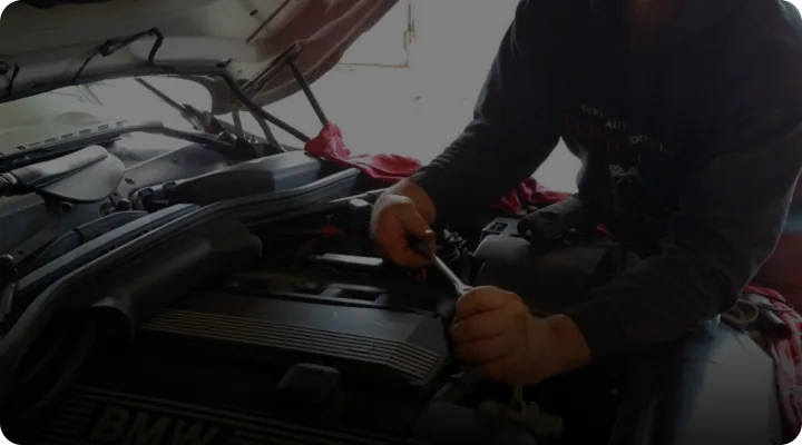 Person working on a BMW car engine with the hood open, using a tool. The individual is focused on the task, wearing a hoodie in a dimly lit garage.