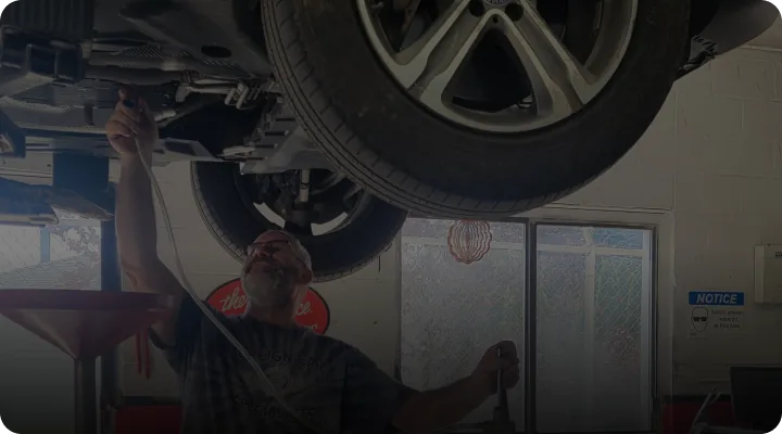 A mechanic works underneath a raised car in a garage. He is turning a wrench on the vehicles underside. A large tire is visible above him. The garage has a window and a notice sign in the background.