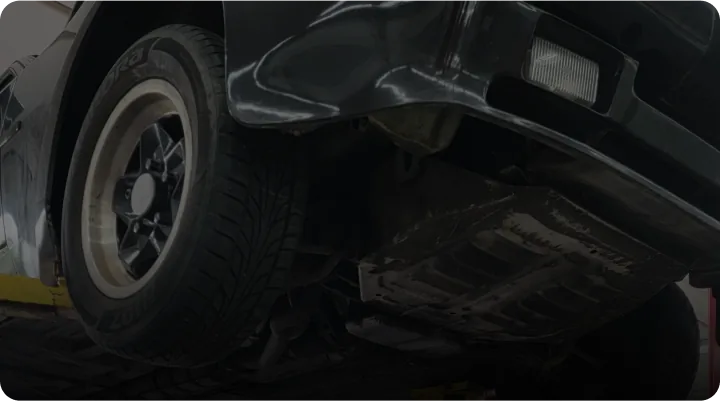 Low-angle view of a black car on a lift in a garage, focusing on the front section with tire and underbody visible. The lighting is dim, emphasizing the cars contours and mechanical details.