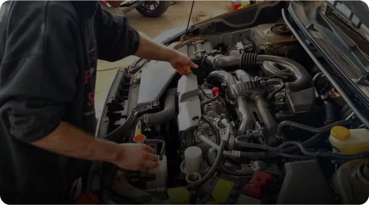 A person in a black sweatshirt works under the hood of a car, handling engine components. Several engine parts and fluid reservoirs are visible, suggesting maintenance or repair is in progress.