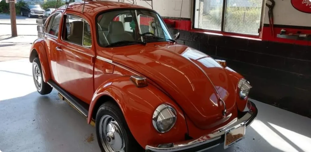 vw repair, auto repair in Poughkeepsie, NY at Foreign Car Specialists. An orange vintage Volkswagen Beetle is parked indoors, with shiny paint reflecting the light. The car is in a garage or workshop, with large windows and a red and black accent wall in the background.