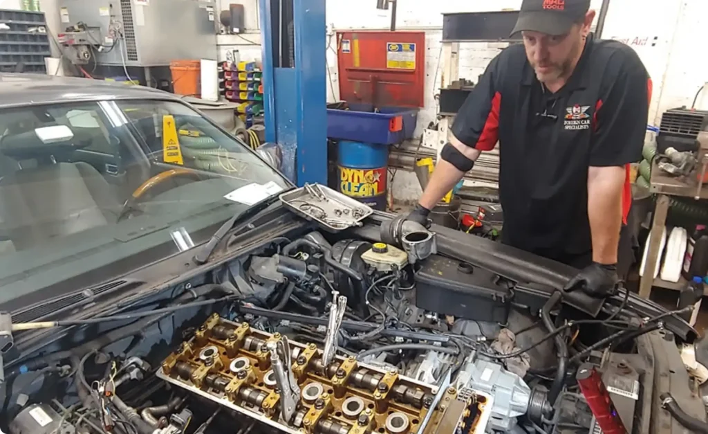 A mechanic wearing a black cap and shirt with red accents inspects a car engine in a garage. The engine hood is open, revealing the internal components. Various tools and equipment are visible in the background.