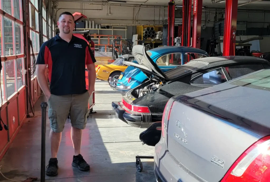 A man stands in a car workshop, wearing a black and red shirt and khaki shorts. Behind him are several cars, including a blue and a yellow one with their trunks open. The garage is spacious with red pillars and ample light.
