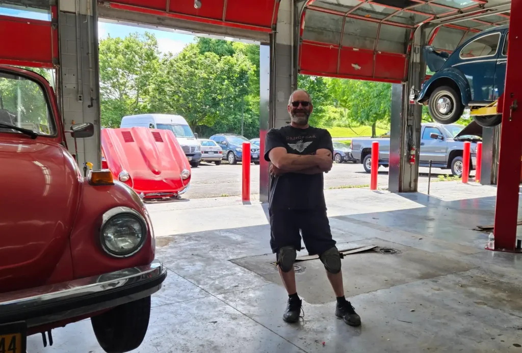A man stands smiling inside an auto repair garage. Hes wearing a black shirt and knee pads. Several cars are visible, including a red car with its hood open and a blue car on a lift. Trees and parked vehicles can be seen outside.
