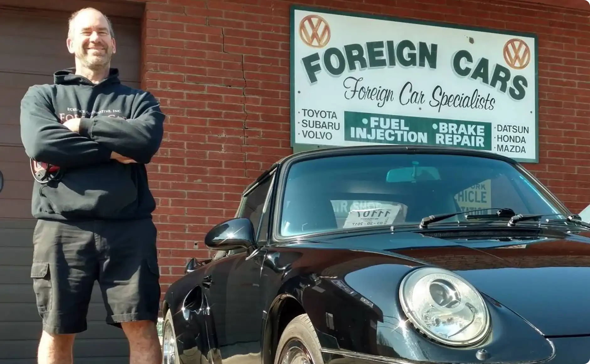 european auto repair, auto repair in Poughkeepsie, NY at Foreign Car Specialists. A person stands confidently with crossed arms next to a black car. The backdrop is a brick building with a sign for Foreign Cars specializing in brands like Toyota and Honda. The person is wearing a black hoodie and shorts.