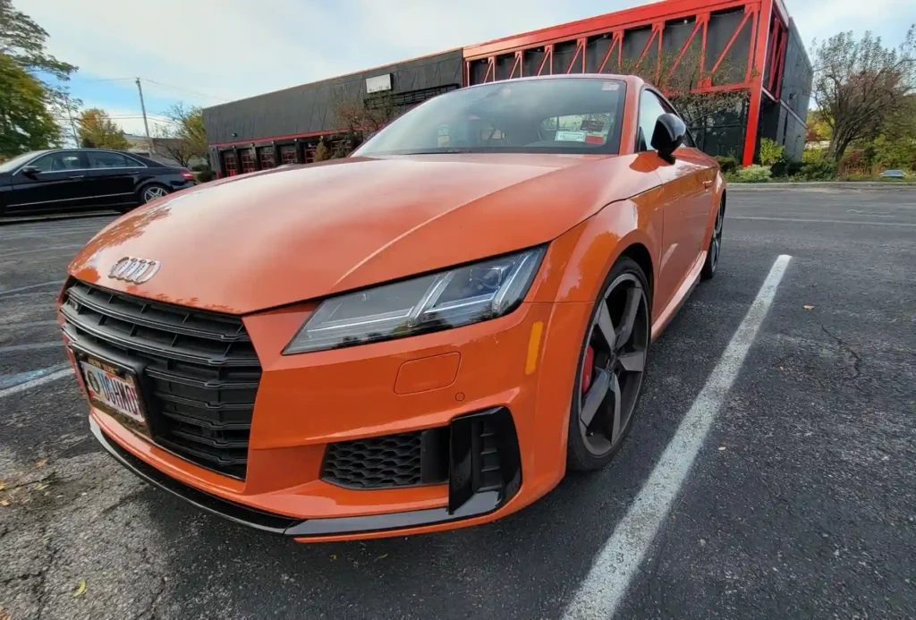 Orange Audi TT sports car parked in a lot with a modern building in the background. The car features sleek lines and sporty details.
