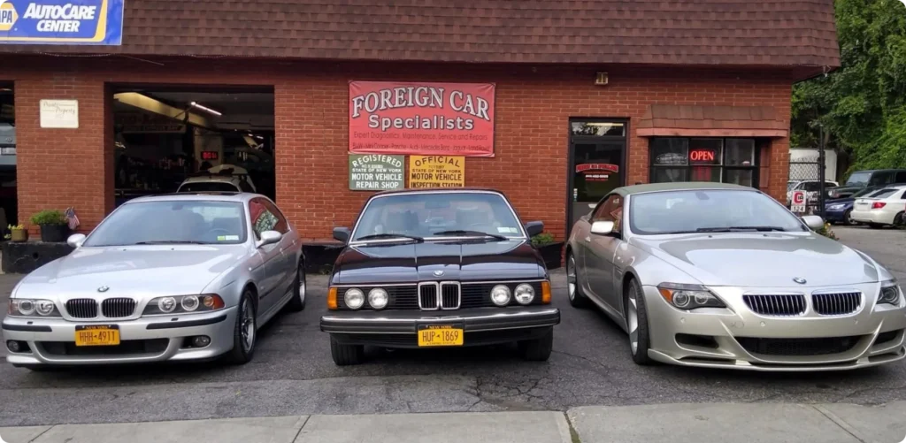bmw repair, auto repair in Poughkeepsie, NY at Foreign Car Specialists. Three BMW cars, including a vintage model, parked in front of a brick building with a Foreign Car Specialists sign. The garage has an Open sign and various automotive signs displayed near the entrance.