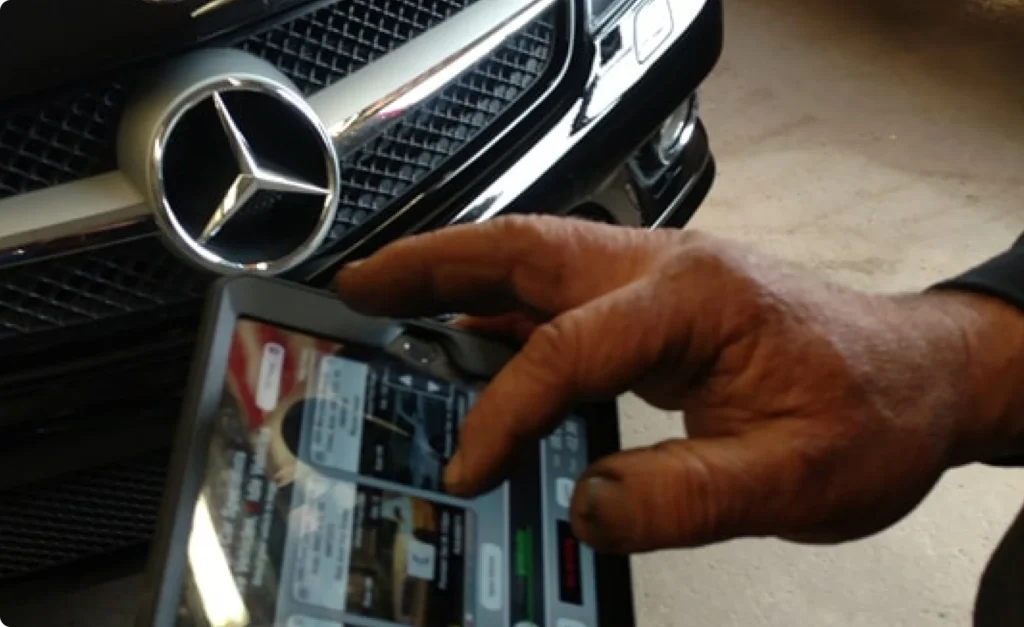engine diagnostics, auto repair in Poughkeepsie, NY at Foreign Car Specialists. A persons hand uses a touchscreen device displaying various icons and information. In the background, the front grille and logo of a black Mercedes-Benz car are visible. The hand is slightly dirty, suggesting recent mechanical work.