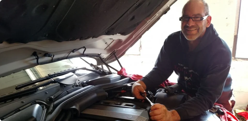 A man wearing glasses and a hoodie is smiling while working under the hood of a car. He is holding a tool, likely repairing or inspecting the engine. The cars hood is open, and sunlight is filtering through a nearby window.