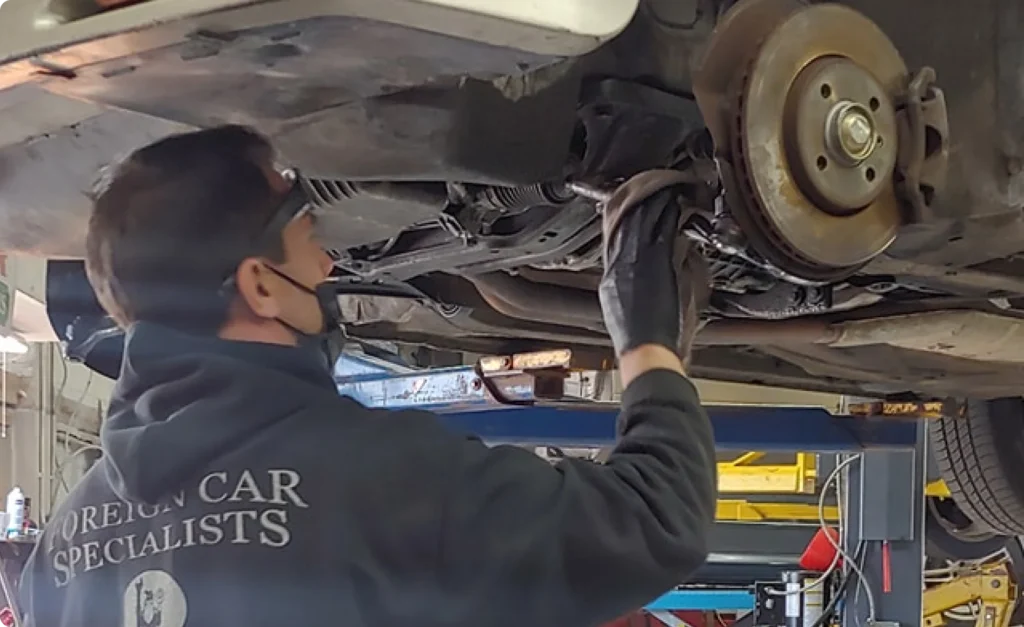 brake repair, auto repair in Poughkeepsie, NY at Foreign Car Specialists. A mechanic wearing a mask and gloves works on the underside of a raised vehicle. The mechanics hoodie reads Foreign Car Specialists. The garage setting includes tools, equipment, and a lift supporting the vehicle.