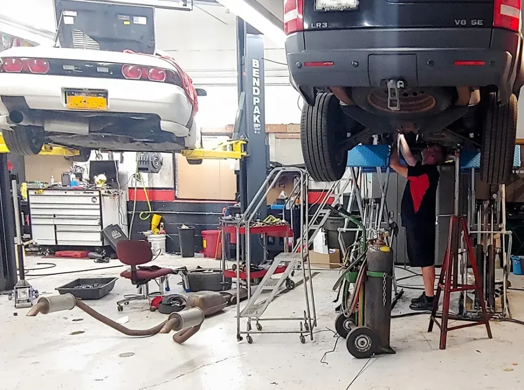 A mechanic works under a black SUV elevated on a lift in an auto repair shop. A white car is on another lift in the background. Car parts are scattered on the floor, and tools are visible on a cluttered workbench.