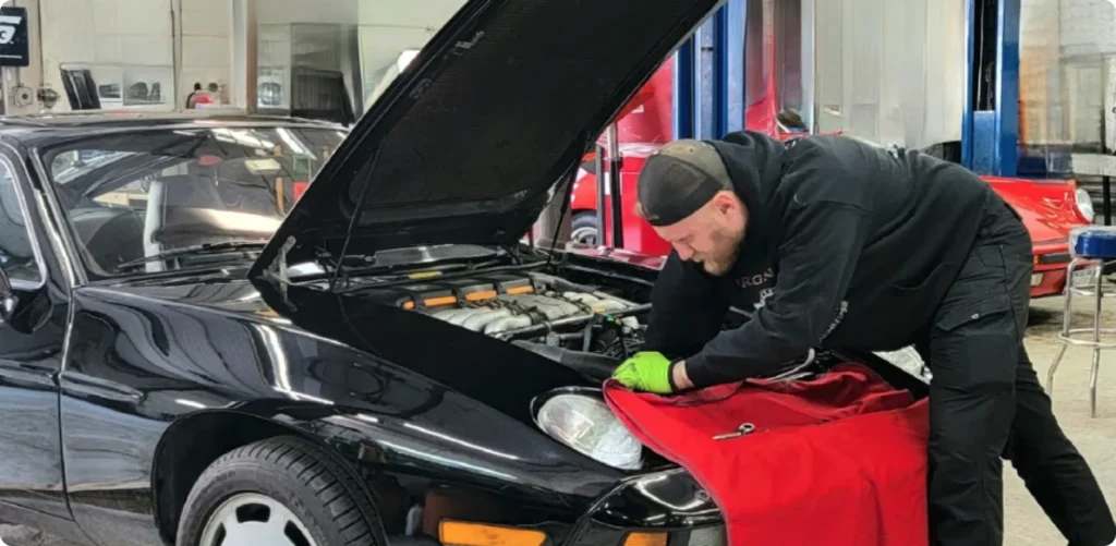 mercedes repair, auto repair in Poughkeepsie, NY at Foreign Car Specialists, A mechanic wearing a black hoodie and green gloves works under the hood of a black car in a garage. The car has a red cover on the front fender. Other vehicles and automotive tools are visible in the background.