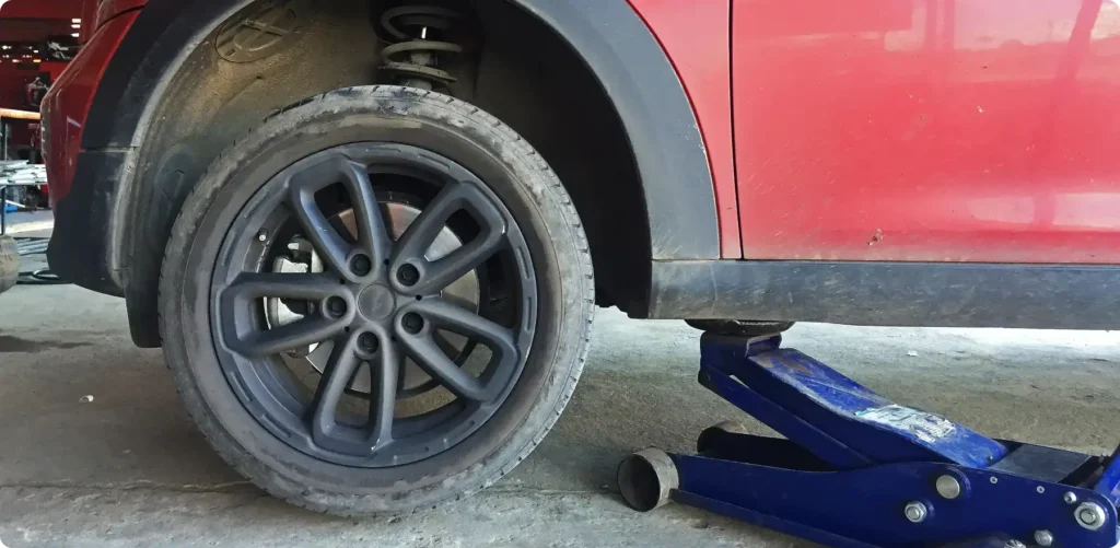 mini repair, auto repair in Poughkeepsie, NY at Foreign Car Specialists. A red car is lifted with a blue hydraulic jack, revealing the rear wheel with black rims in a garage. The suspension system and part of the undercarriage are visible on a concrete floor.