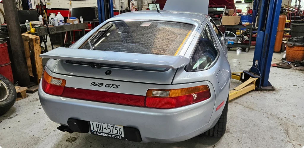 porsche repair, auto repair in Poughkeepsie, NY at Foreign Car Specialists. A silver Porsche 928 GTS is positioned on a car lift inside a garage. The rear hatch is open, and various tools and equipment are visible in the background, indicating a maintenance or repair setting.