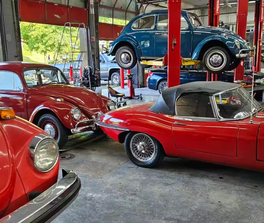 walkway location, auto repair in Poughkeepsie, NY at Foreign Car Specialists. A garage scene with classic cars: a blue Volkswagen Beetle is on a lift, another red Beetle is parked nearby, and a red convertible with wire wheels is in the foreground. The space is well-lit and organized.