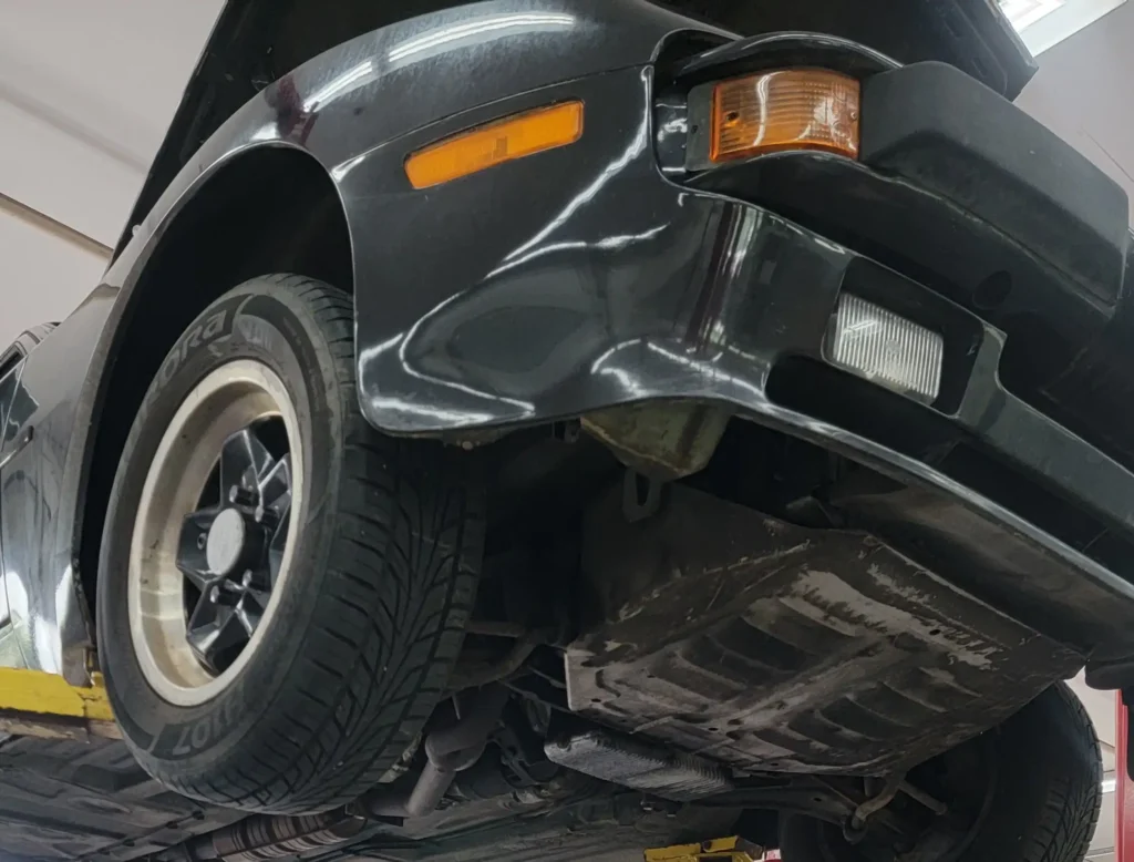 tire services, auto repair in Poughkeepsie, NY at Foreign Car Specialists. Close-up view of a black car lifted on a mechanics lift. The focus is on the front passenger side, showing the tire, undercarriage, headlights, and open hood. The vehicle appears to be in a garage for maintenance or inspection.