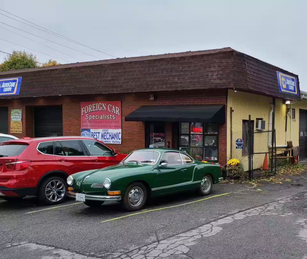 south campus location, auto repair in Poughkeepsie, NY at Foreign Car Specialists. A small auto repair shop with several garage bays is depicted. Two classic cars, a green convertible and a blue vintage sedan, are parked outside. A red SUV is parked to their left. Signs for Foreign Car Specialists are visible on the building.